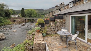 Une terrasse en pierre avec une table et des chaises, donnant sur une rivière, avec un bâtiment en pierre et un pont en arrière-plan.