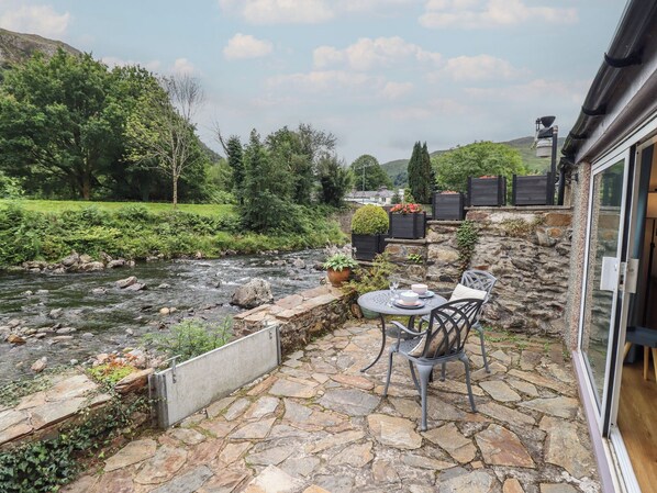 Une terrasse en pierre avec une table et des chaises, donnant sur une rivière, et entourée de verdure et de murs de pierre.