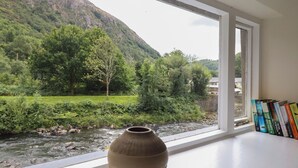 Une pièce dotée d’une grande fenêtre donnant sur une rivière et la verdure environnante, un vase posé sur le rebord de la fenêtre et une bibliothèque remplie de livres.