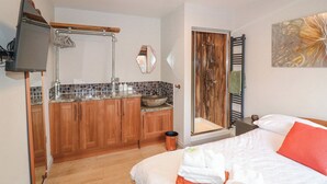 Une chambre d’hôtel compacte dotée d’une kitchenette, d’une douche avec une porte en verre, d’un lit avec une literie blanche et orange, et d’un miroir fixé au mur.