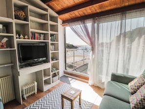 Una sala de estar con televisor, estanterías, un sofá y vista al mar.