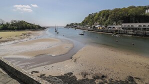 A coastal scene with a sandy beach, calm water, boats, and a stone wall.