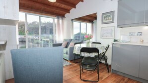 A cozy living room with a blue sofa, a dining table with chairs, and a kitchen area with cabinets and a window overlooking a garden.