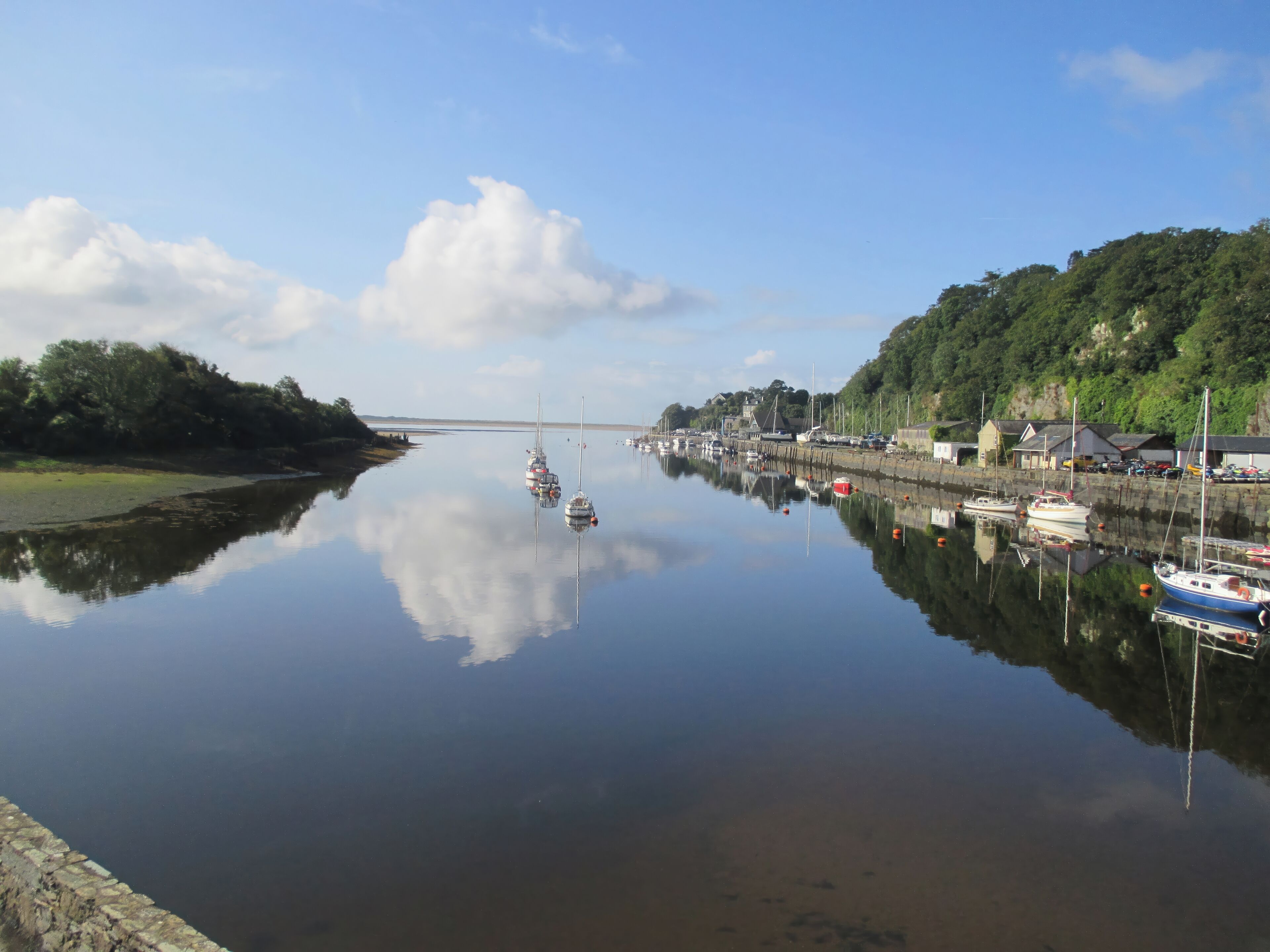 A calm harbor with boats docked along the quay, surrounded by greenery and buildings.