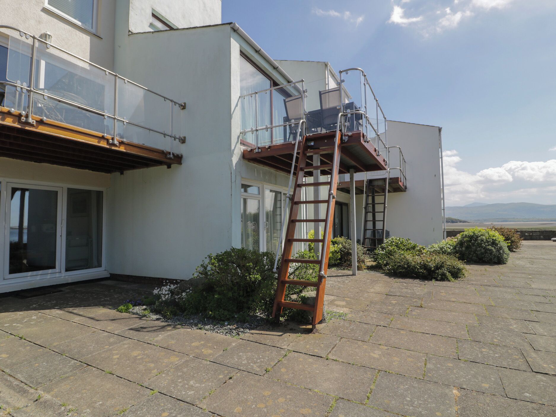 A modern building with a glass balcony, a wooden ladder, and a paved courtyard.