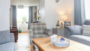 Un salon moderne avec un canapé, un fauteuil et une table basse en bois. On y trouve une lampe sur une table d’appoint, ainsi qu  ’une fenêtre avec des rideaux.