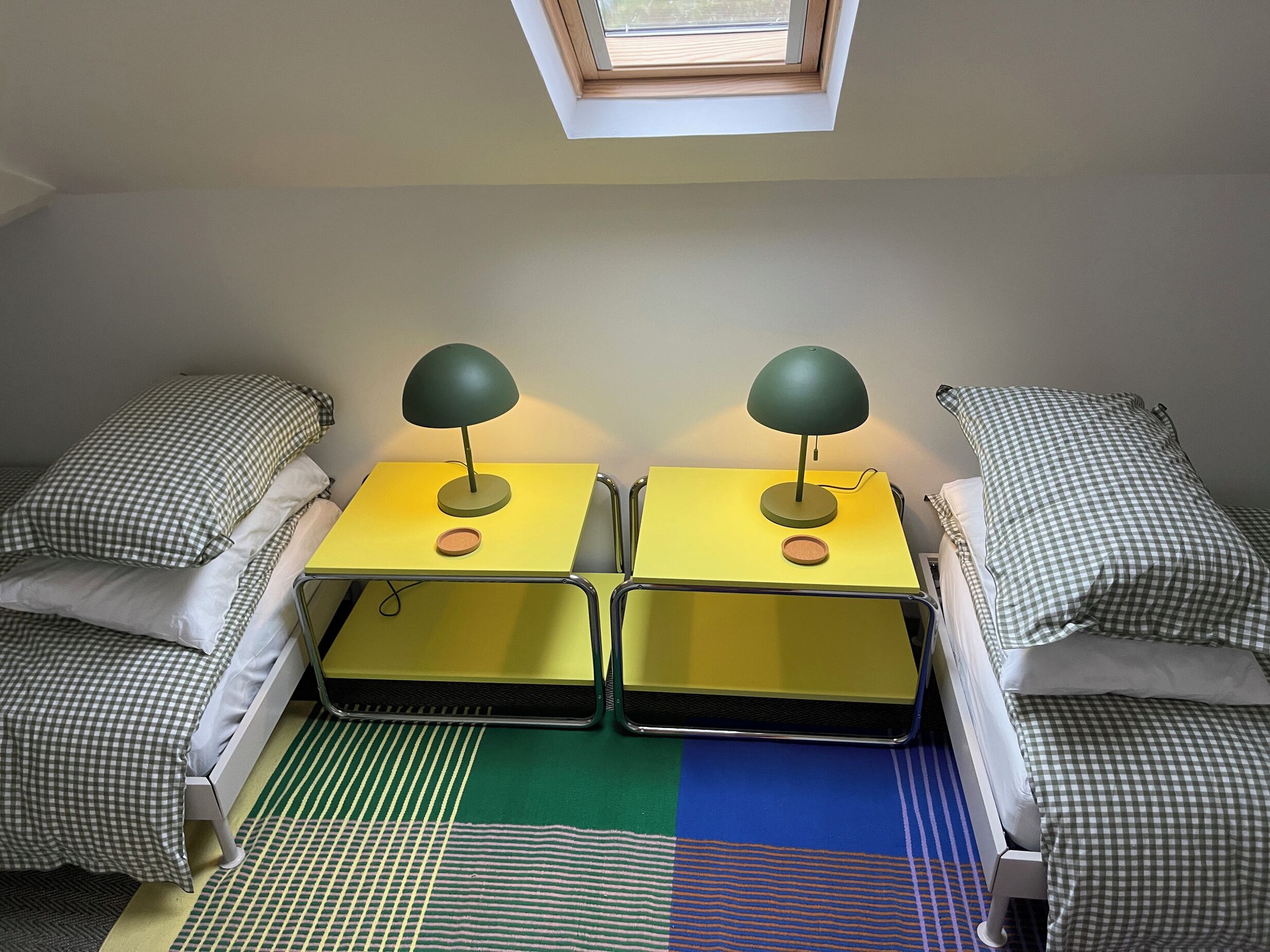 A room with two beds, each with a checkered pillow and a green lamp on a yellow nightstand. There is a skylight above the beds.