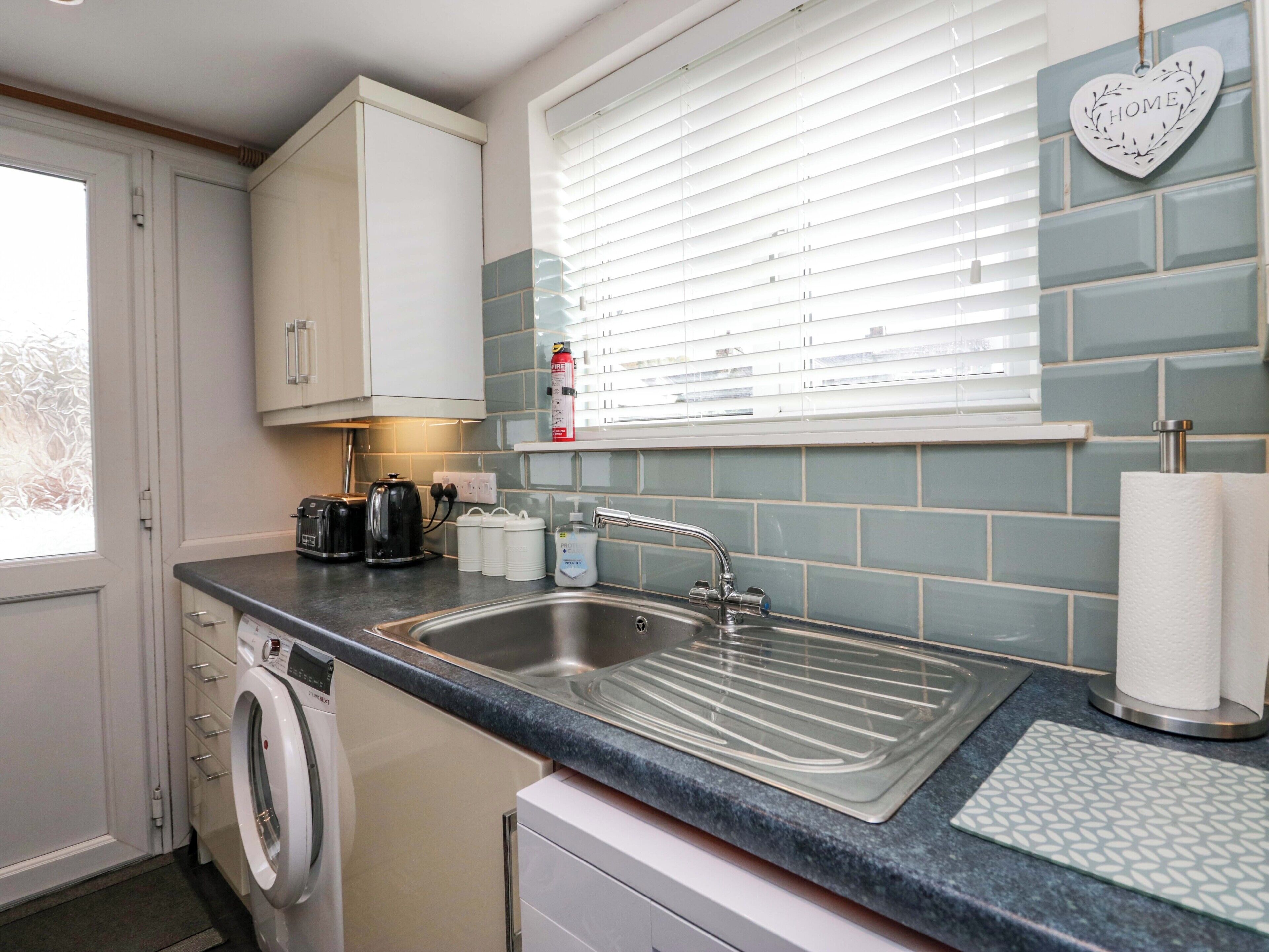 A modern kitchen with a stainless steel sink, white cabinets, a washing machine, and a window with blinds.