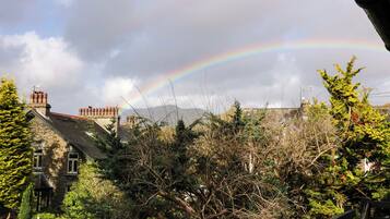 A stone building with a sloped roof and multiple chimneys, surrounded by dense greenery and a rainbow in the sky.