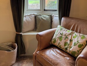 A leather armchair with a leaf-patterned pillow, a window seat with a view of greenery, and a woven basket.
