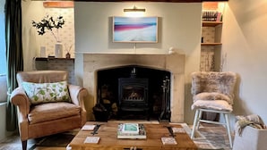 A cozy living room with a fireplace, leather armchairs, a coffee table with books, and a painting above the mantel. - Meadow View Cottage (Cheltenham)