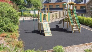 A playground with swings and a slide, surrounded by greenery and a building with a sloped roof. - 4 The Manor House Hillfield Village (Dartmouth)