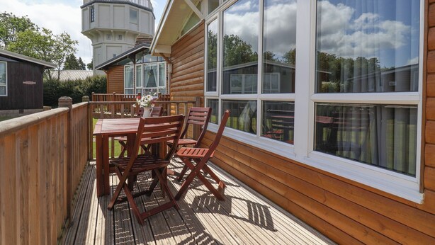 A wooden deck with a table and chairs, large windows, and a small building with a flag. - Pebble Beach Chalet (Seaton)