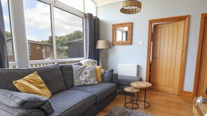 A cozy living room with a grey sofa, a small round table, a grey rug, a wooden door, and a window with a view of buildings outside. - Pebble Beach Chalet (Seaton)