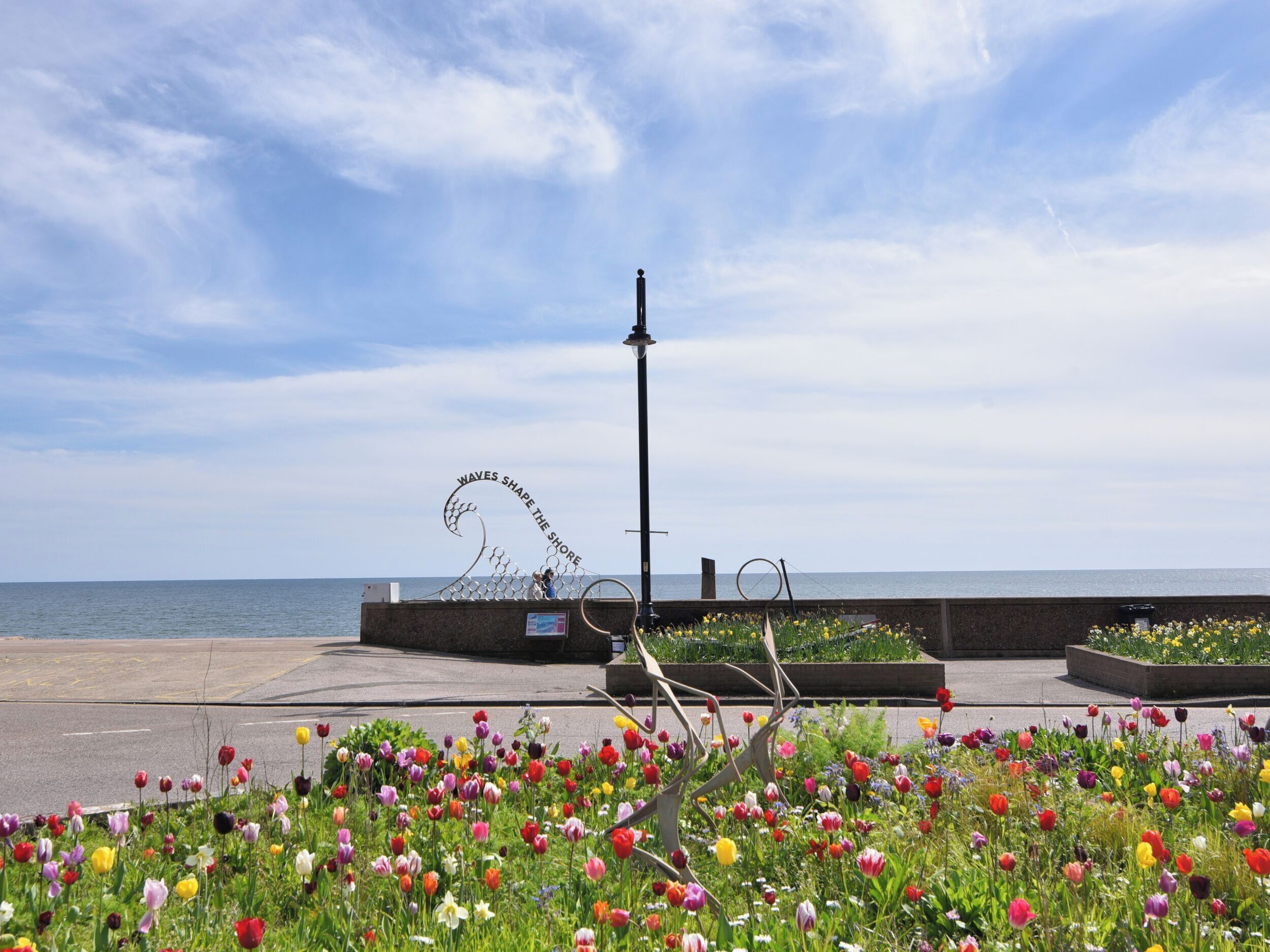 A coastal promenade with a metal sculpture, flower beds, and a view of the sea.