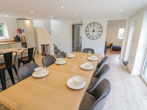 A dining area with a long wooden table set for six, surrounded by chairs. A large wall clock is visible, and there is a staircase leading to another level.
