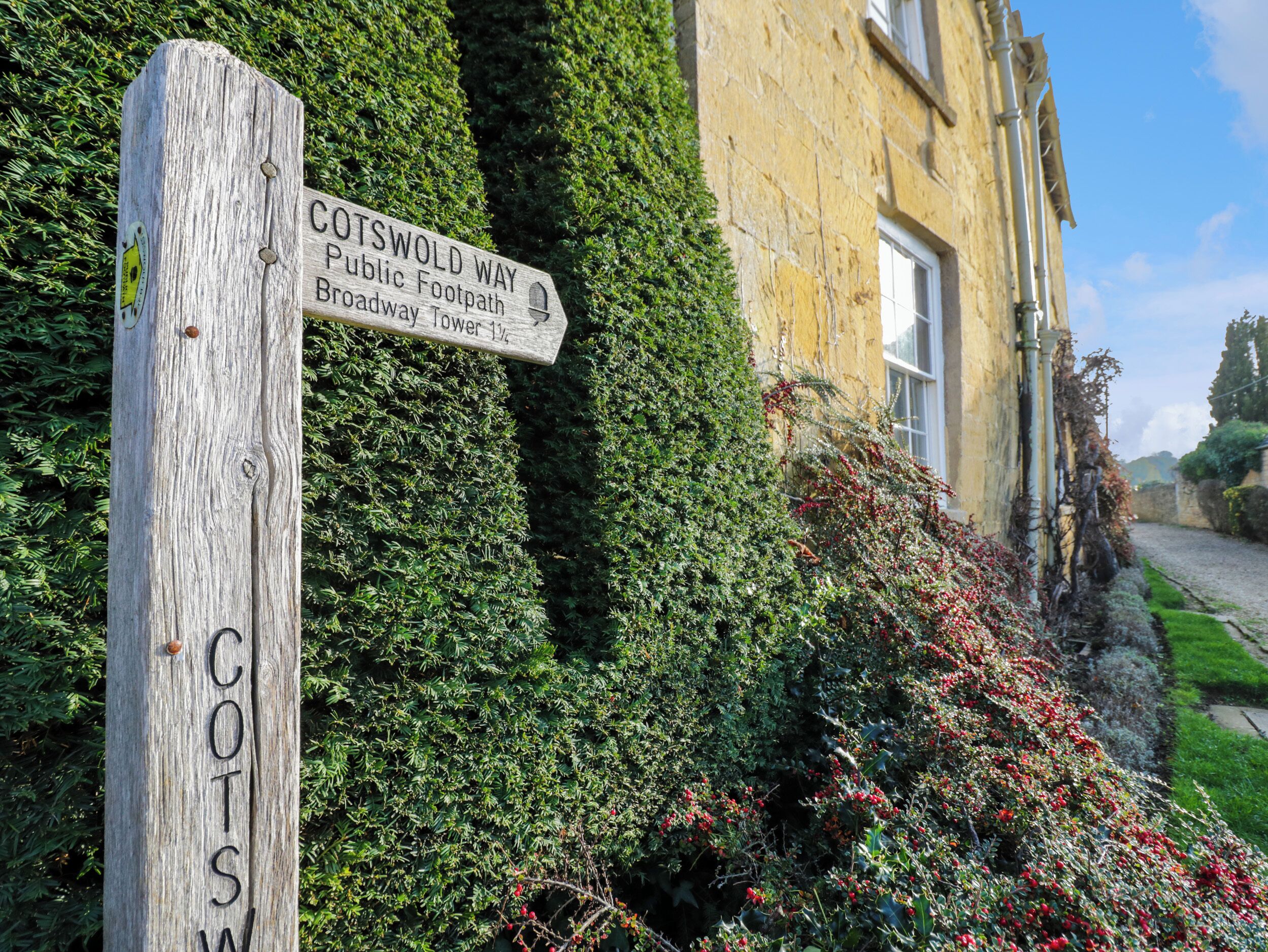 Un cartel de madera con 'COTSWOLD WAY' y 'Broadway Tower 1½' escrito, sobre un fondo de un edificio y abundante vegetación.
