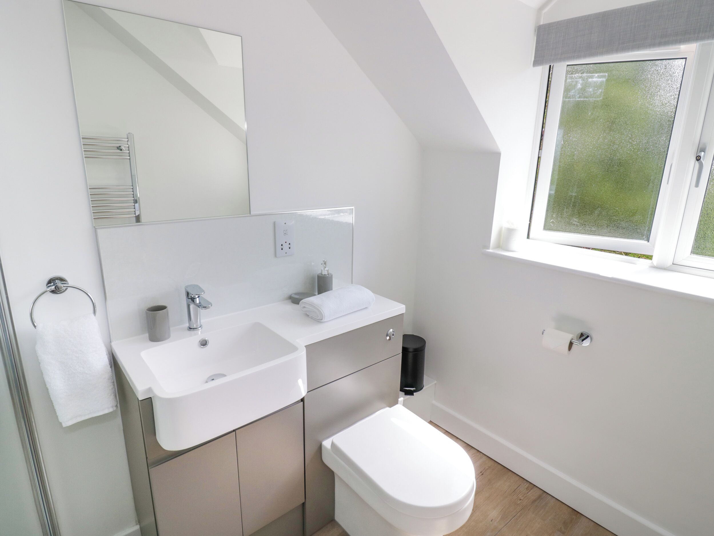 Een moderne badkamer met een wit wastafel, spiegel en toilet. Er is een raam met uitzicht op groen buiten.