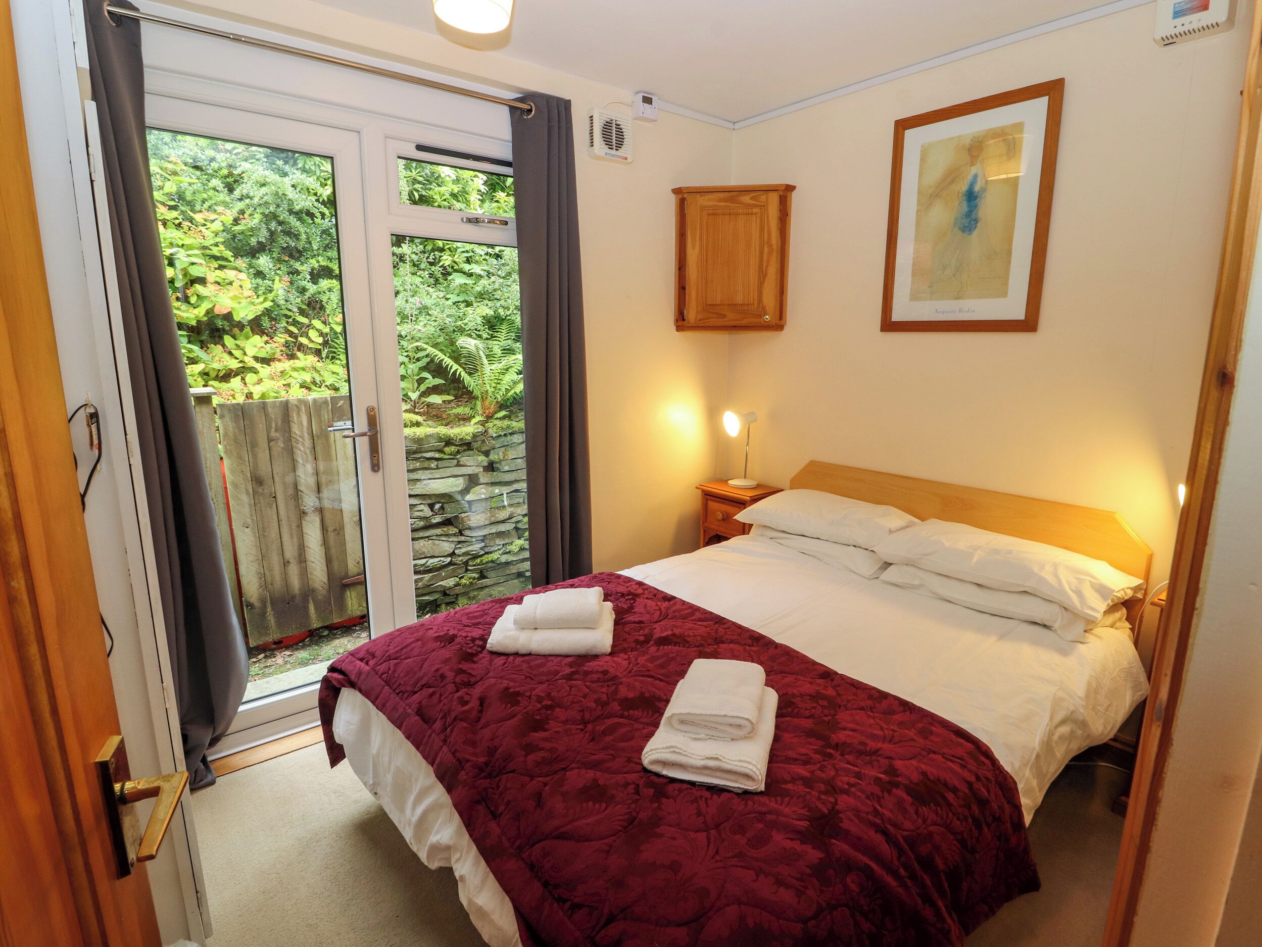 A bedroom with a large bed, a nightstand, a bedside lamp, a framed picture on the wall, and a sliding glass door leading to a garden.