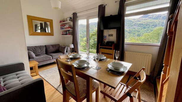 A dining area with a table set for four, a sofa, a bookshelf, and a view of the outdoors.