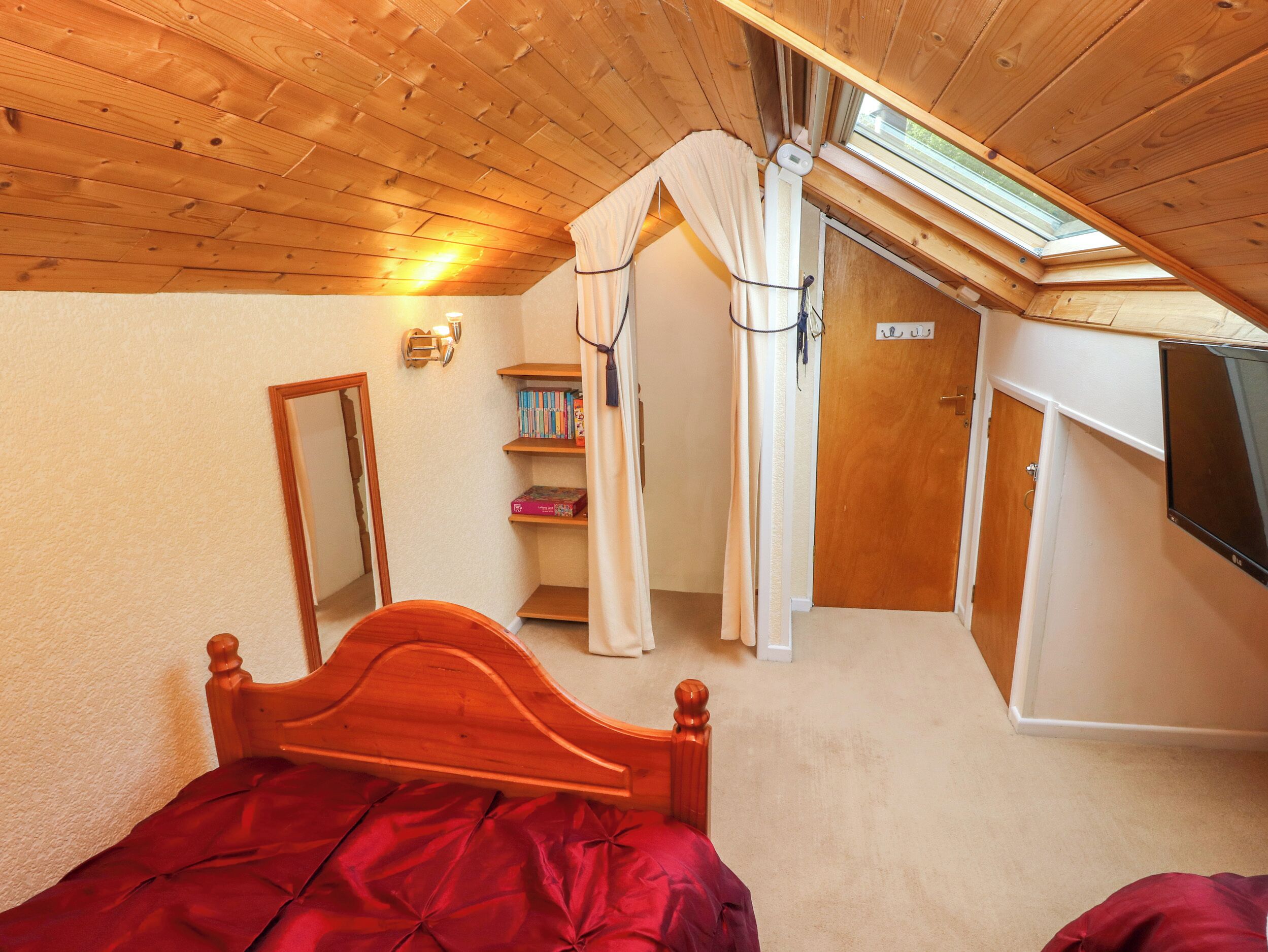 A bedroom with a wooden ceiling, a bed, a skylight, a bookshelf, and a door.