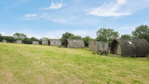 A row of rustic, wooden cabins with pitched roofs, arranged on a grassy hillside. - Sheep Shed @ Penbugle Organic Farm (Liskeard)