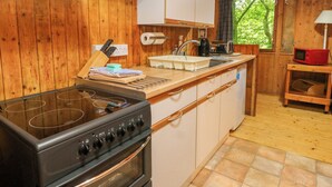 A compact kitchen with wooden cabinets, a stove, a sink, and a window with a view of trees. - Lodge 3 - "beech" (Isle of Arran)