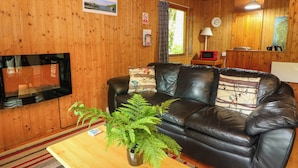 A wooden interior with a leather sofa, a television, a dining area, and a window with curtains. - Lodge 3 - "beech" (Isle of Arran)