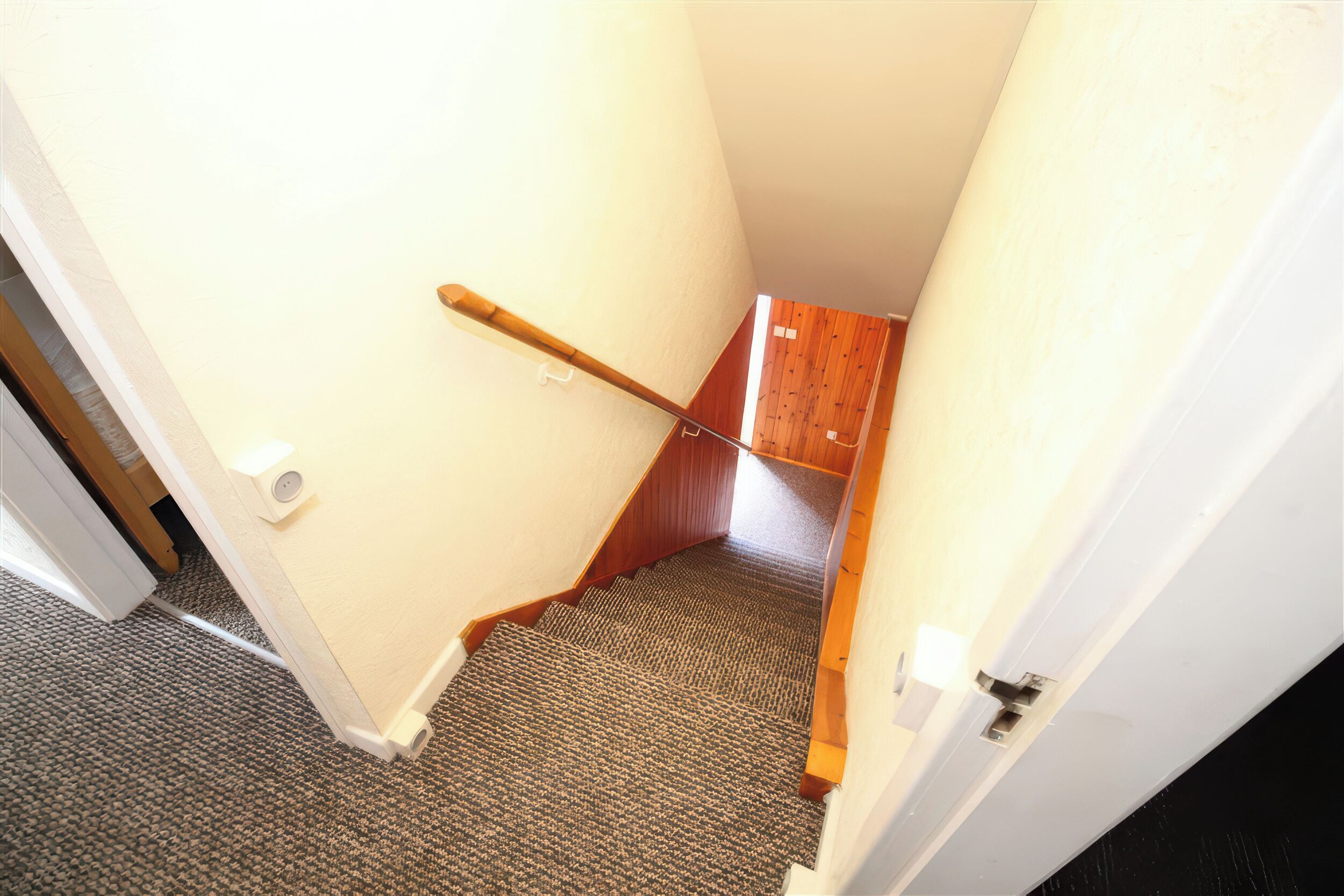A carpeted stairway with a wooden handrail, white walls, and a door leading to another room.