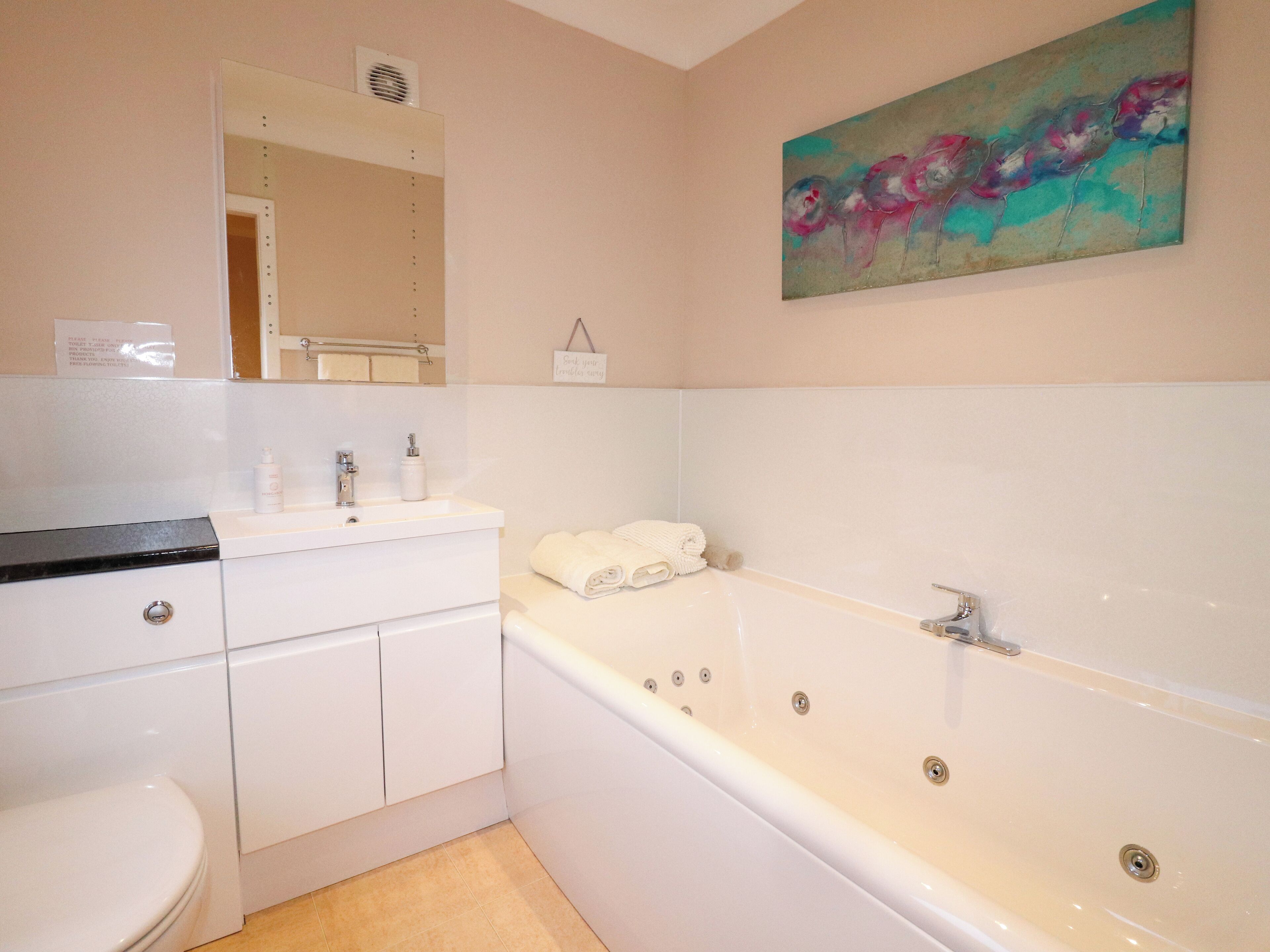 A modern bathroom with a white sink, toilet, and bathtub. There is a mirror above the sink and a painting on the wall.