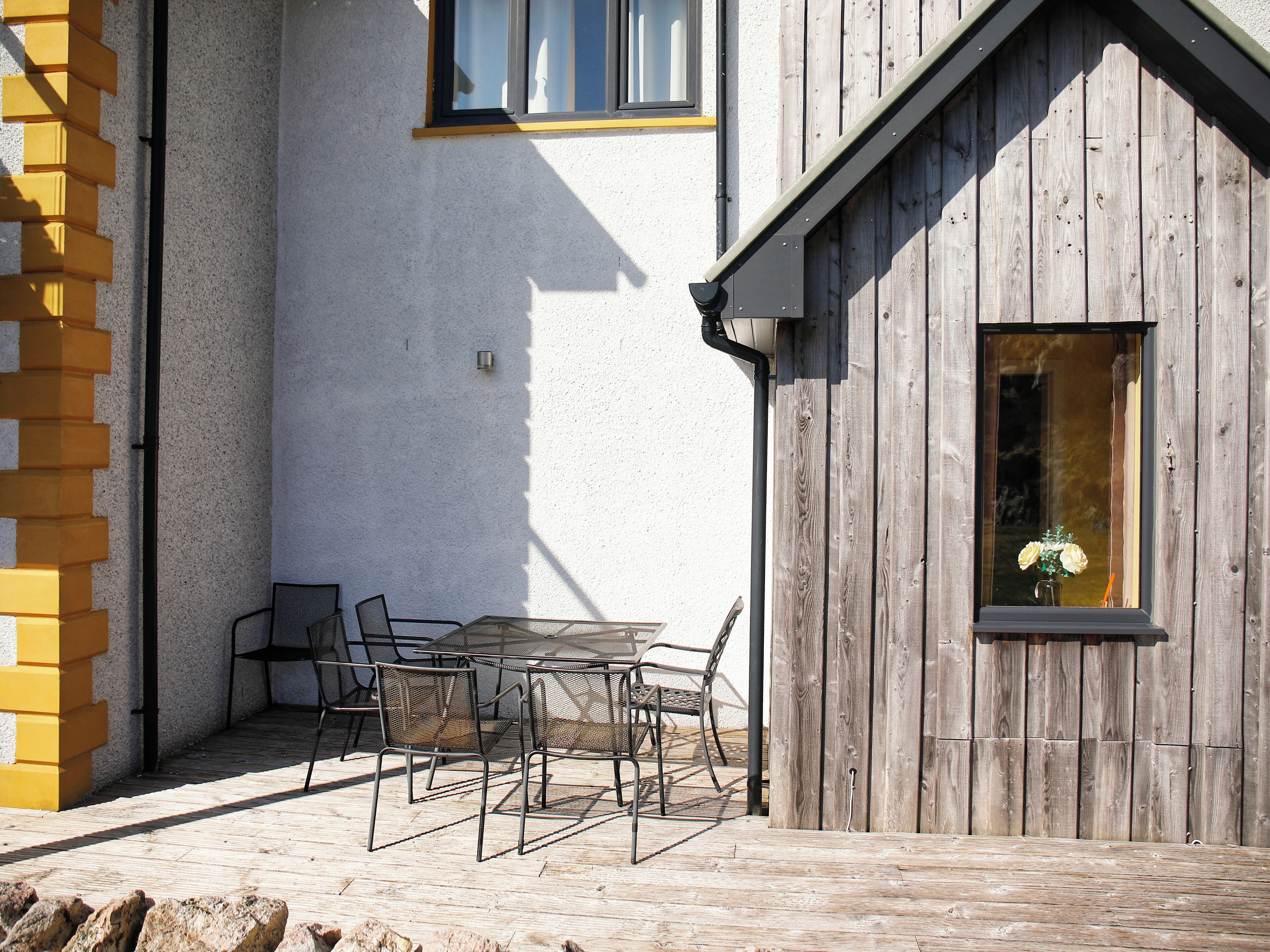 Une terrasse extérieure avec une table et des chaises, une clôture en bois et une fenêtre ornée d’un vase de fleurs.