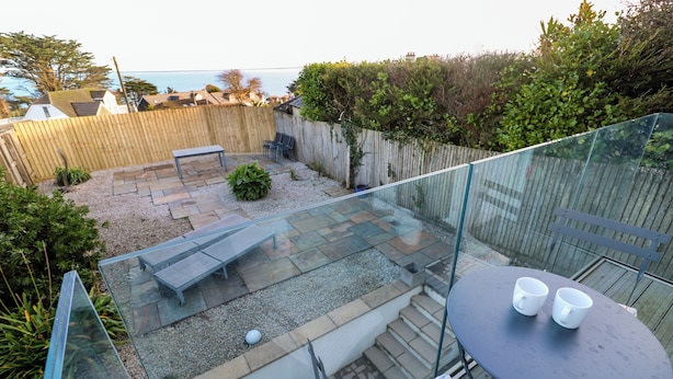 A glass-enclosed balcony with a table and chairs, a small garden area with a bench, and a view of the sea. - Azur (St Ives)