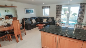 A modern living room with a black leather sofa, wooden dining table, and a kitchen area with wooden cabinets and a black countertop. - Azur (St Ives)