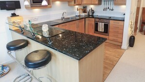 A modern kitchen with wooden cabinets, a granite countertop, a stainless steel oven, and a glass bar with stools. - Azur (St Ives)