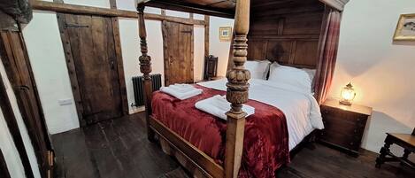 Een traditioneel hemelbed met een rode sprei en witte lakens, een houten vloer en een nachtkastje met een lantaarn.