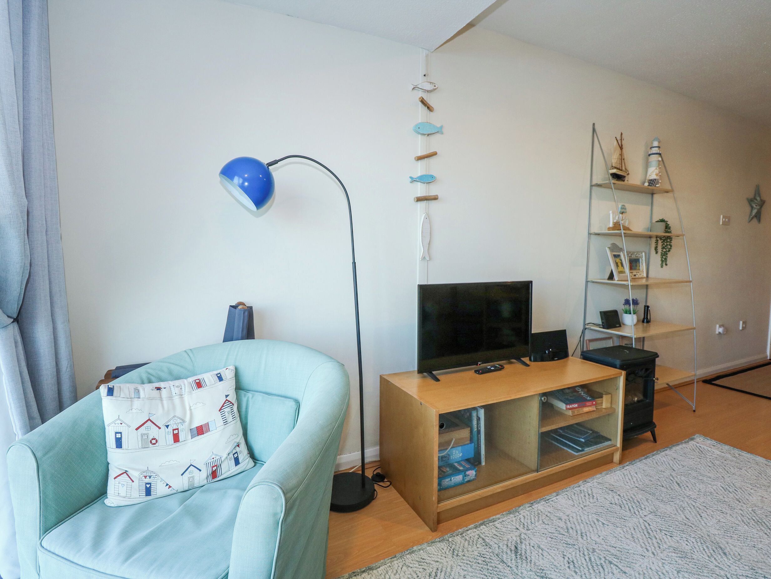 A living room with a light blue armchair, a blue floor lamp, a TV on a wooden stand, and a bookshelf with decorative items.