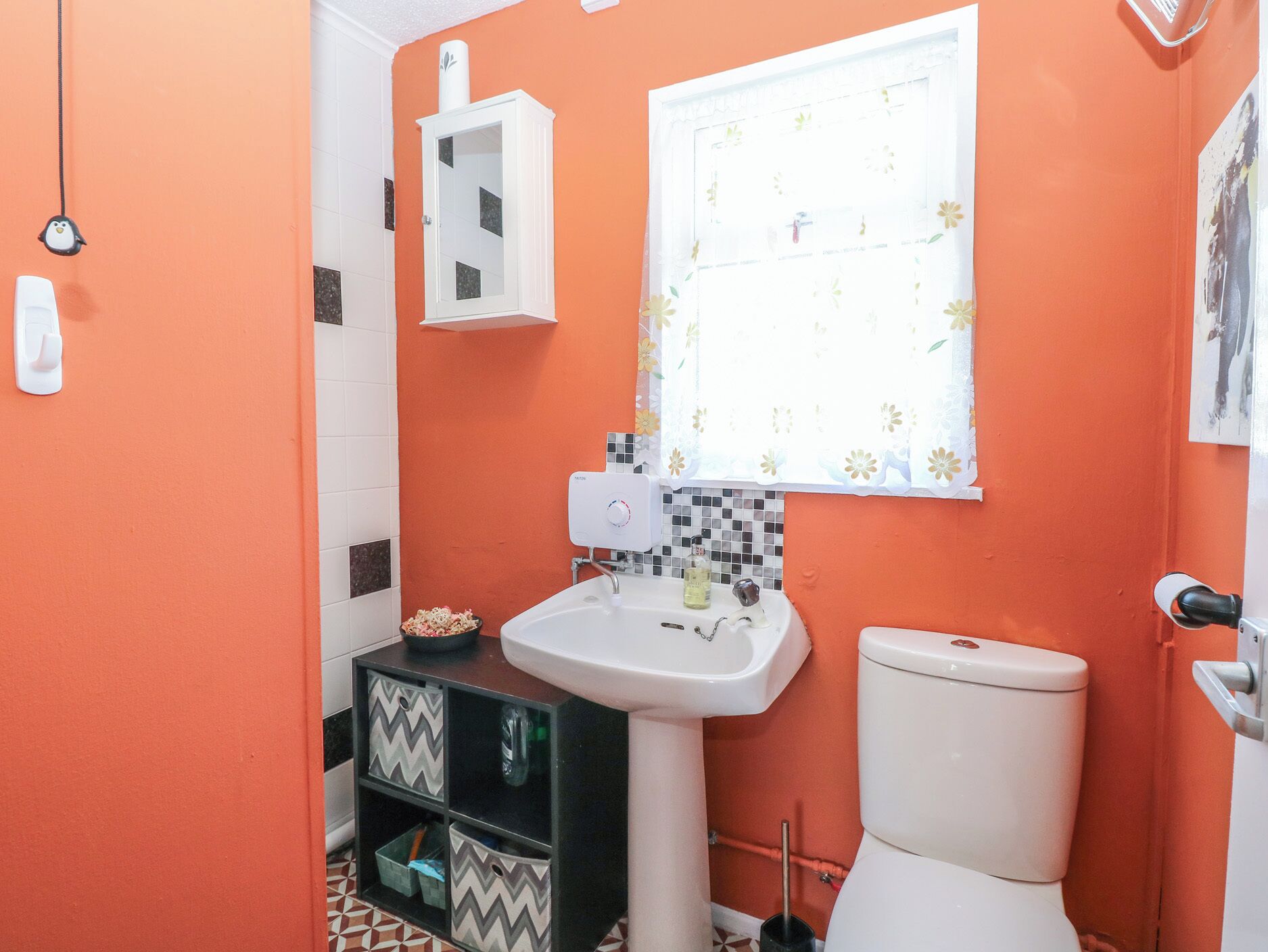 A bathroom with an orange wall, a white sink, a toilet, and a window with floral curtains.