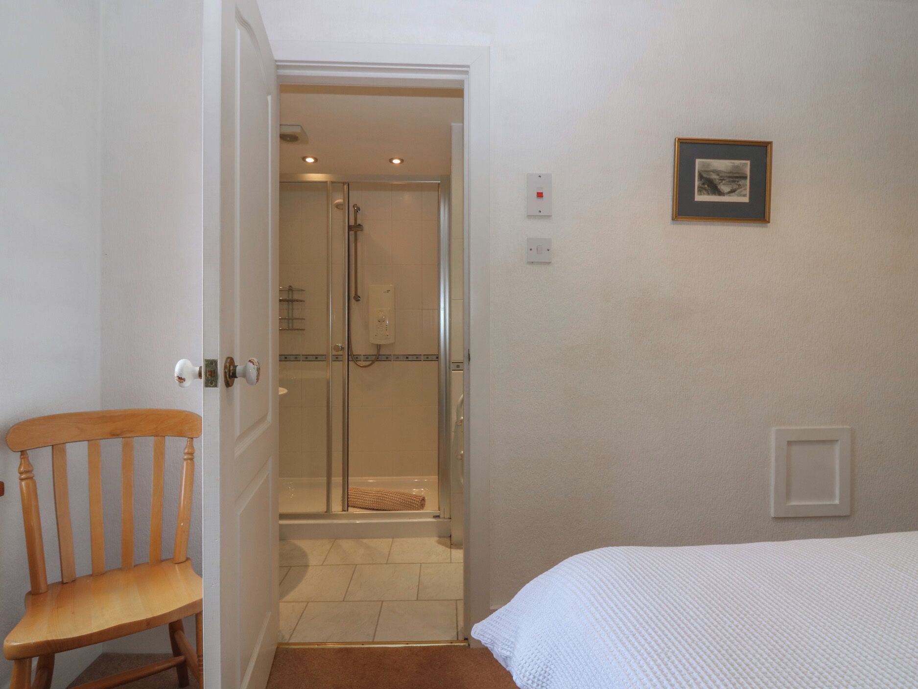 A bedroom with a wooden chair, a bed, a framed picture on the wall, and a shower area with a glass door.