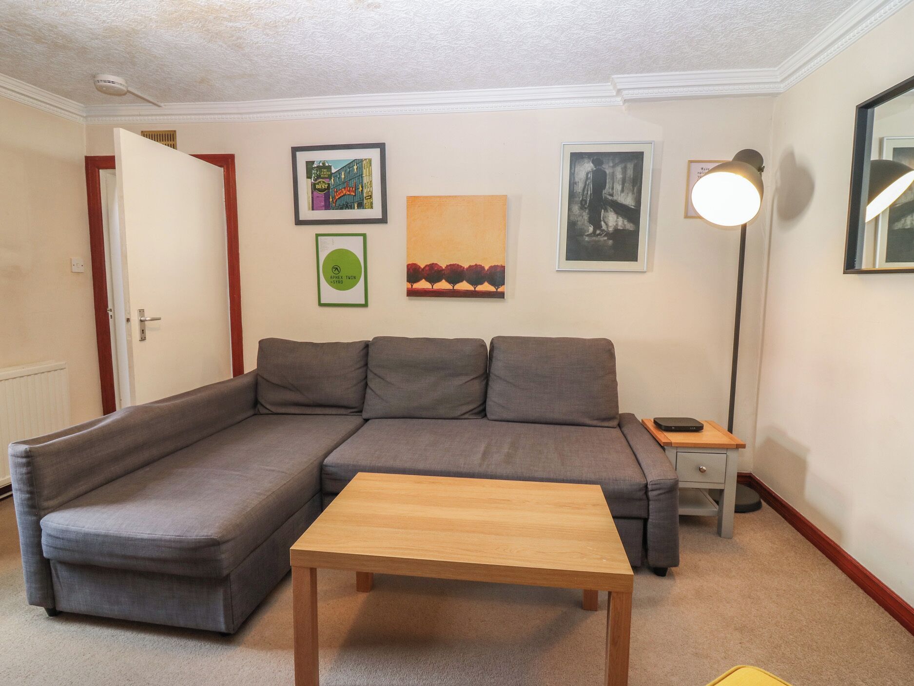 A living room with a gray sectional sofa, a wooden coffee table, a floor lamp, and framed artwork on the walls.