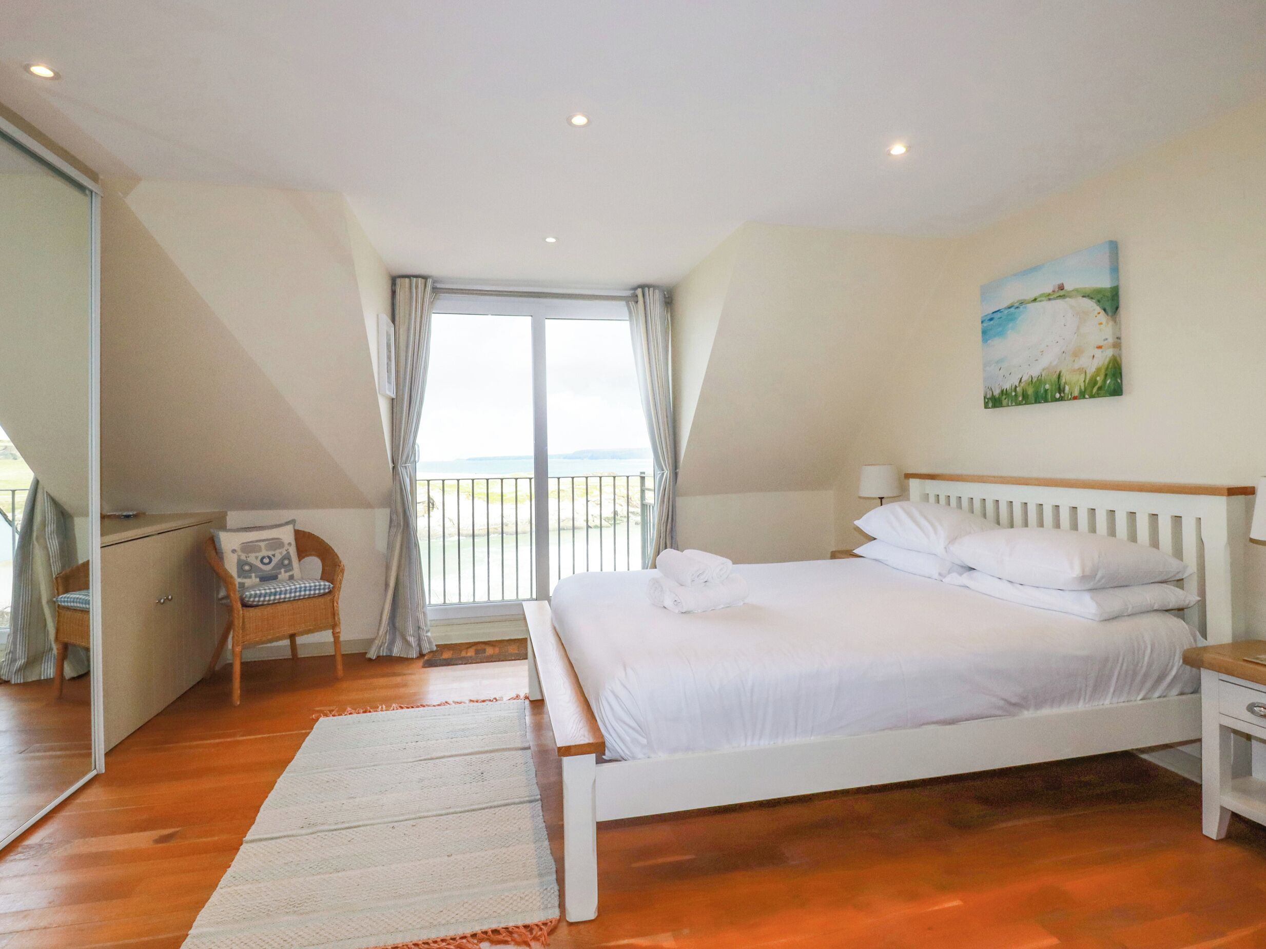 A bedroom with a large bed, a chair, a desk, and a view of the sea through a balcony.