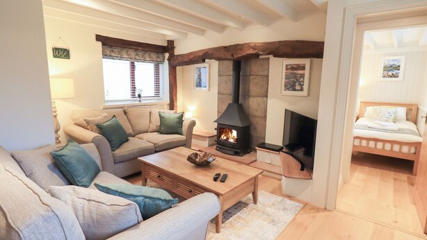 A cozy living room with a sofa, a coffee table, a fireplace, and a bed in the adjacent room. - Jasmine Cottage (Keswick)