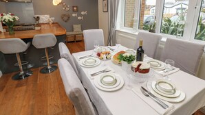 A dining area with a table set for four, a kitchen in the background, and a view of the outside through large windows. - Seafarer's Rest (Chichester)
