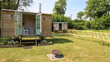 A rustic wooden cabin with an open door, a chimney, and a small outdoor area with a wheelbarrow and a garden bed.