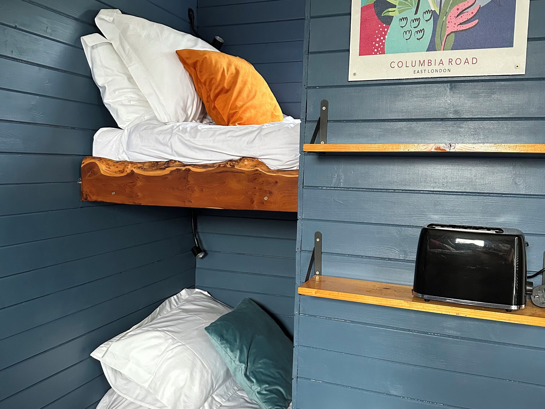 Een stapelbed met witte en oranje kussens, een zwarte tas op een plank, en een poster van COLUMBIA ROAD in Oost-Londen.
