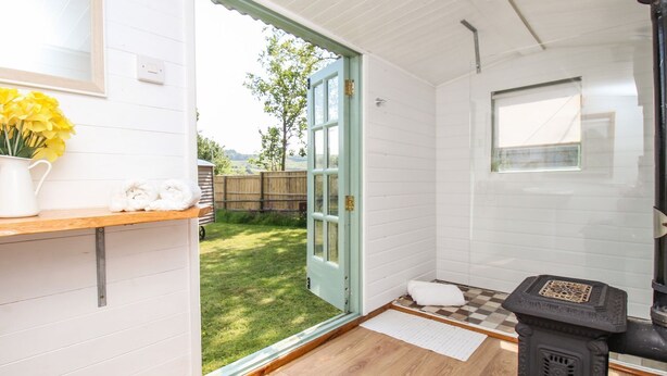 A cozy room with a wooden floor, a white wall, a small window, and a black stove. - Acres Meadow (Dorchester)