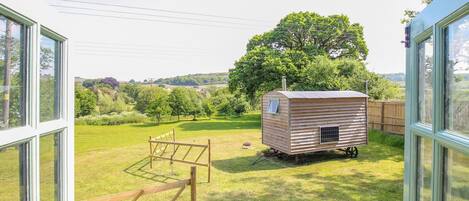 A wooden structure in a grassy yard with a view of a tree and hills in the background.