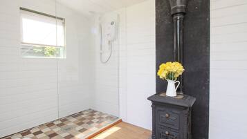 A bathroom with a shower, a white tiled wall, a checkered floor, and a black wood-burning stove with a vase of yellow flowers.