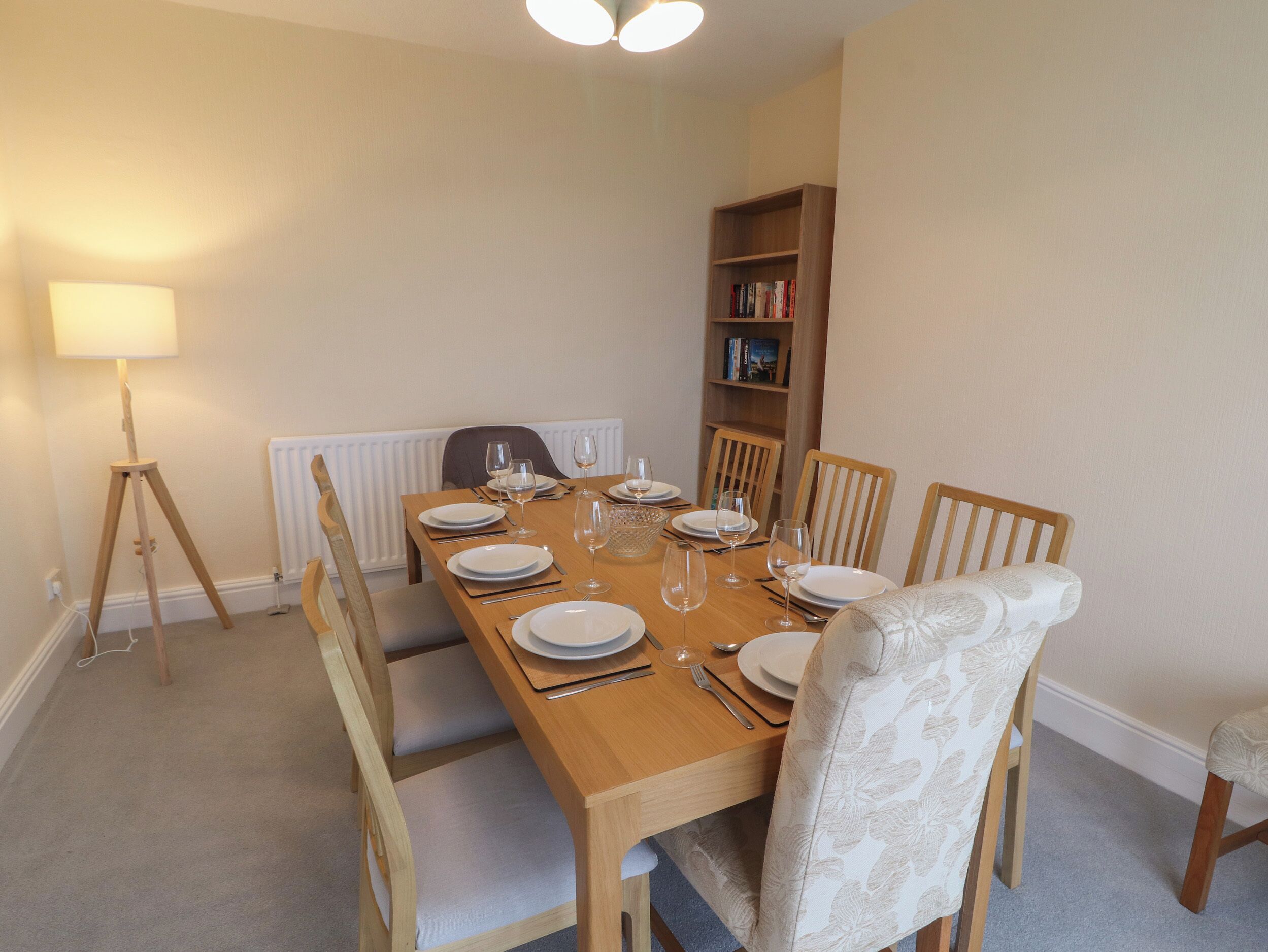 Comedor con mesa de madera dispuesta para seis personas, rodeada por seis sillas. Se observa una estantería contra la pared y una lámpara de pie a la izquierda.