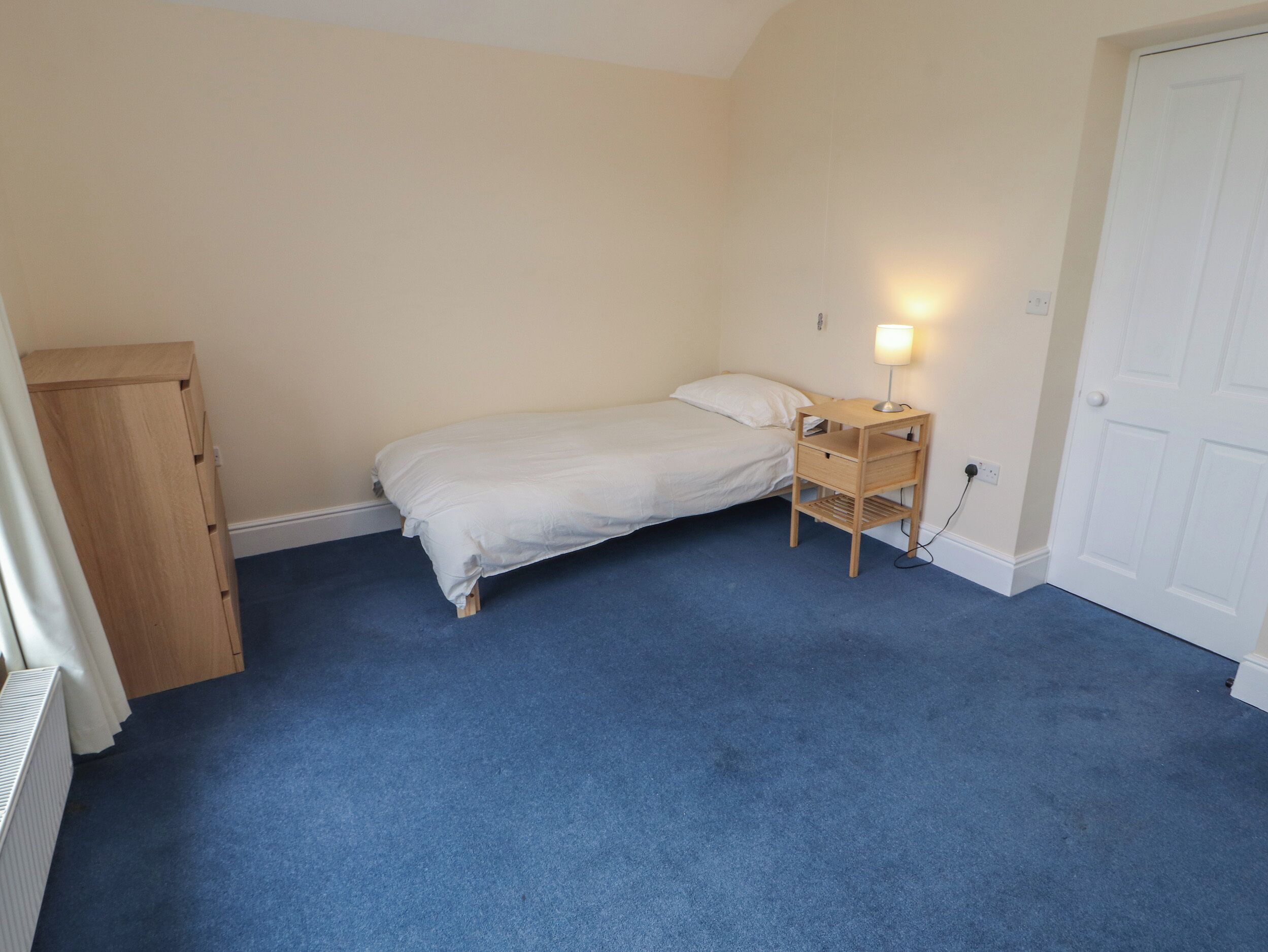 A bedroom with a single bed, a wooden nightstand with a lamp, a radiator, and a door.
