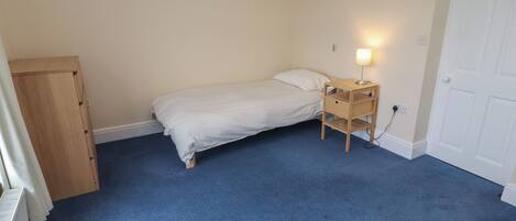 A bedroom with a single bed, a wooden nightstand with a lamp, a radiator, and a door.